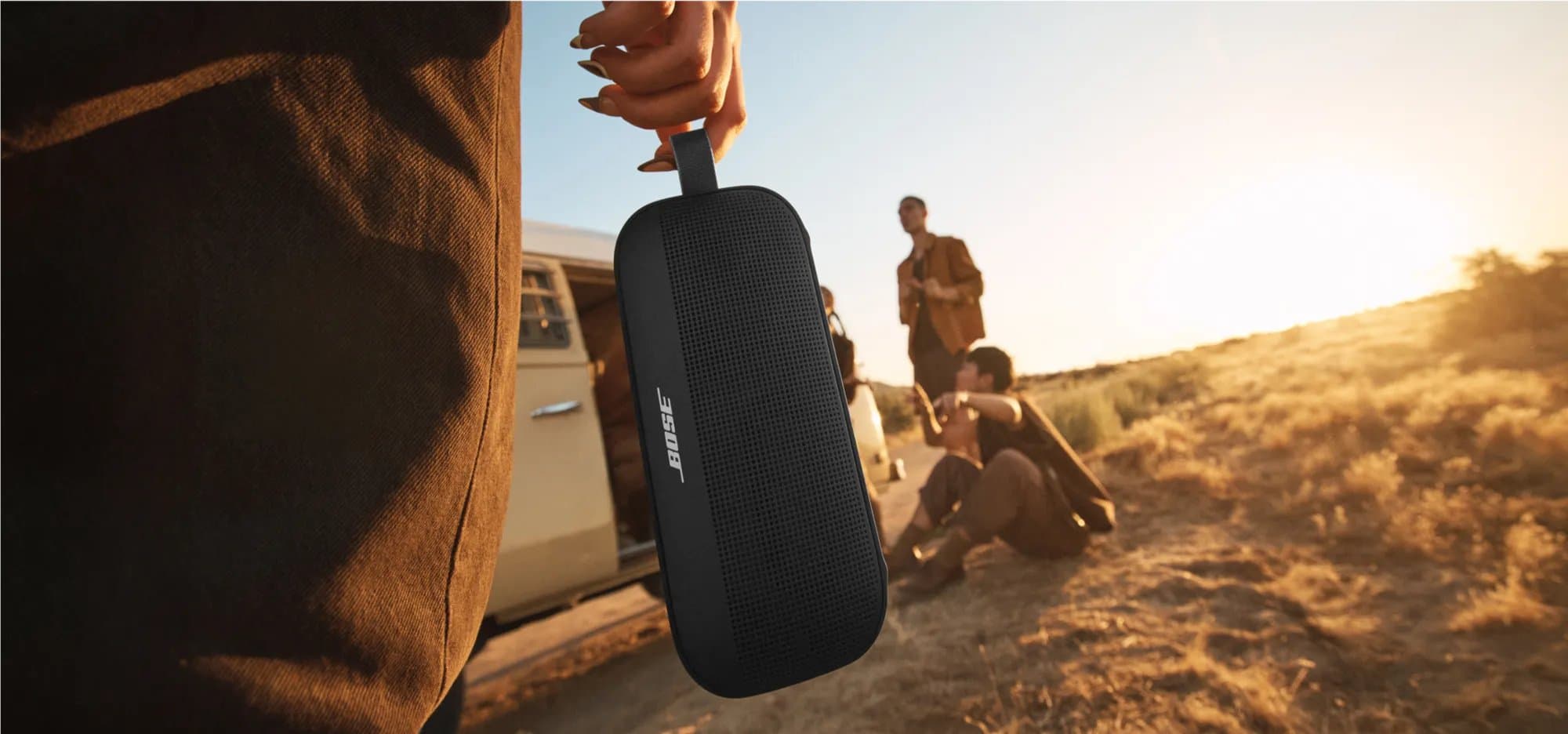 Black Bose SoundLink Flex (2nd Gen) carried by its strap to friends relaxing beside a camper van in warm evening light